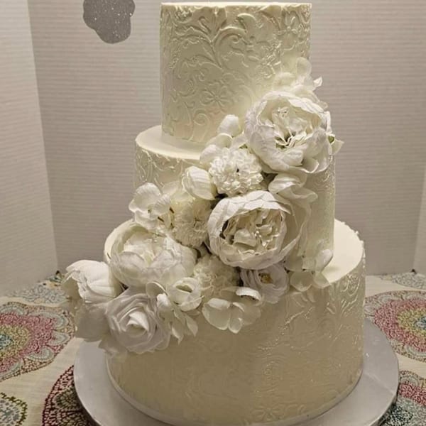 A three-tiered white wedding cake with intricate floral patterns and white sugar flowers.