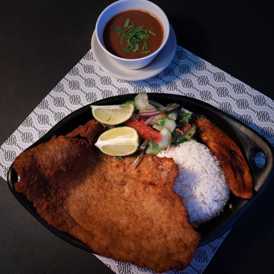 A Colombian platter with breaded pork, rice, salad, plantain, lime, and a side of red bean soup.