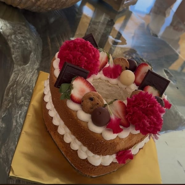 Heart-shaped cake with white frosting, strawberries, chocolate, cookies, and pink flowers on a gold base.