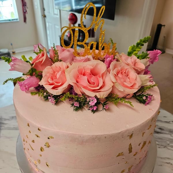 Pink cake with gold flakes, topped with pink roses, greenery, and a gold "Oh Baby" sign.
