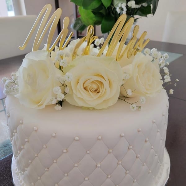 White wedding cake with quilted fondant, white roses, baby's breath, and a gold "Mr & Mrs" topper.
